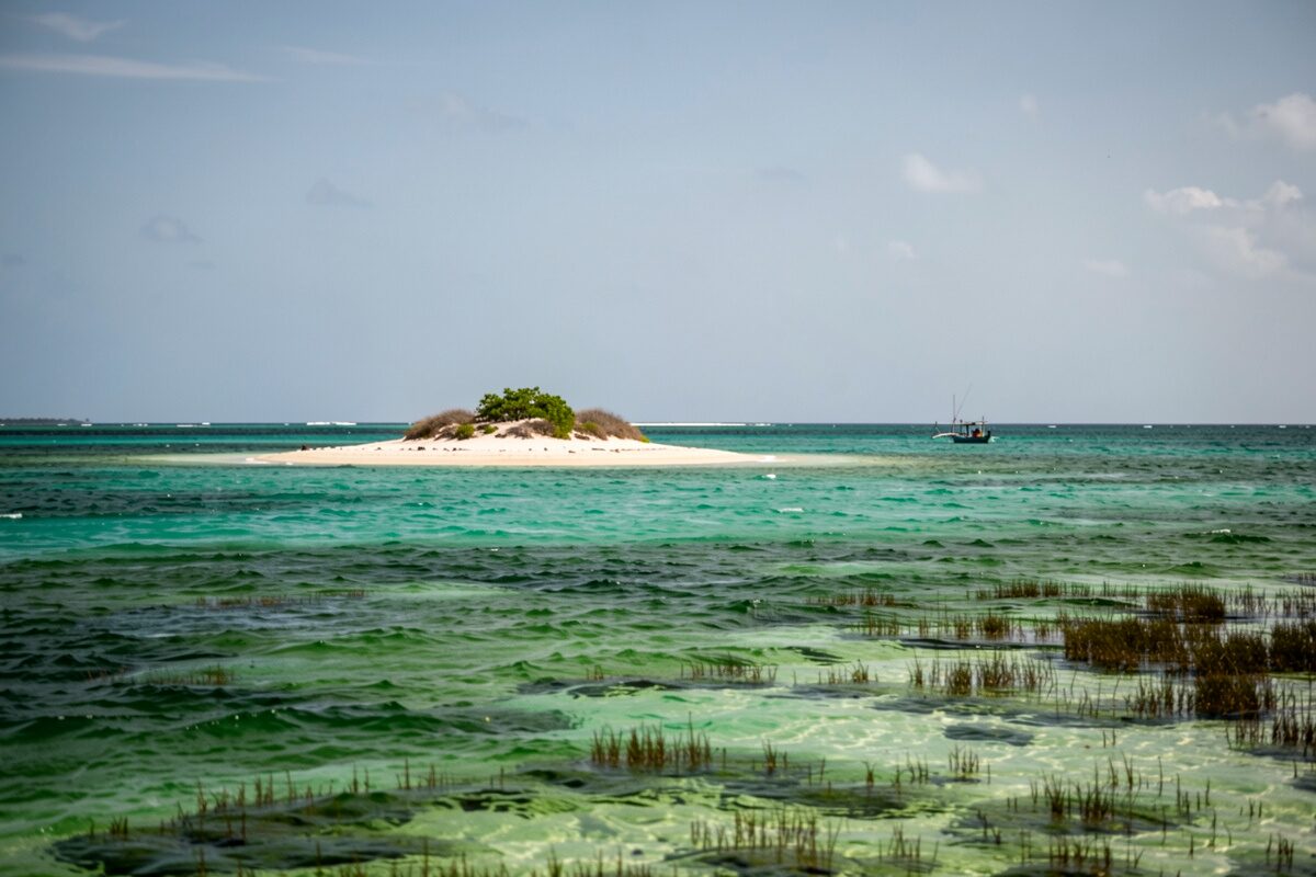 Un lagon d'eau vert émeraude dégradée, tombants sombres sous l'eau, îlots bas et barque de pêche au loin, ciel large.